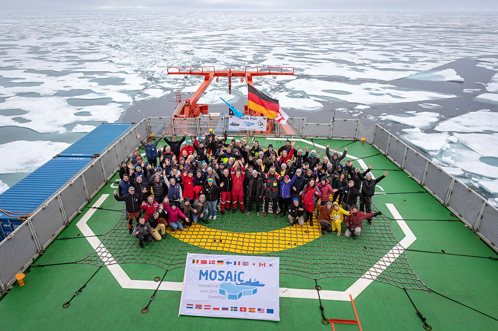 High angle group shot of the MOSAiC Cruise participants waving on the deck of the polarstern with sea ice in the background